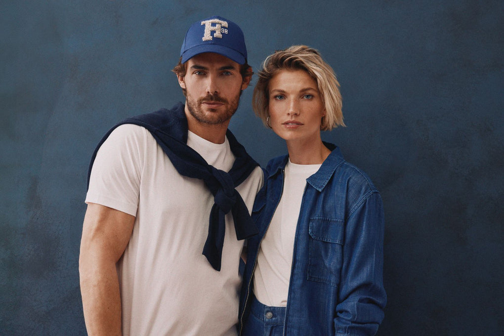 Mann mit blauem Cap, weißem T-Shirt und dunkelblauem Pullover über den Schultern steht neben Frau in Jeanshemd und weißem Shirt – beide blicken frontal in die Kamera.
